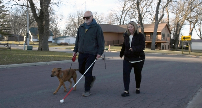 Jim Baker walking down a paved street with his white cane, dog, and partner Susan