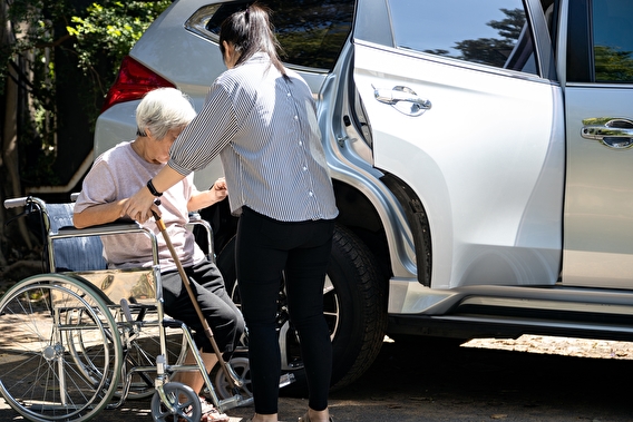 A person helping an eldery individual out of a wheelchair and into the back seat of an SUV