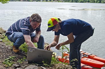 a man and a woman looking at a computer on the ground next to river